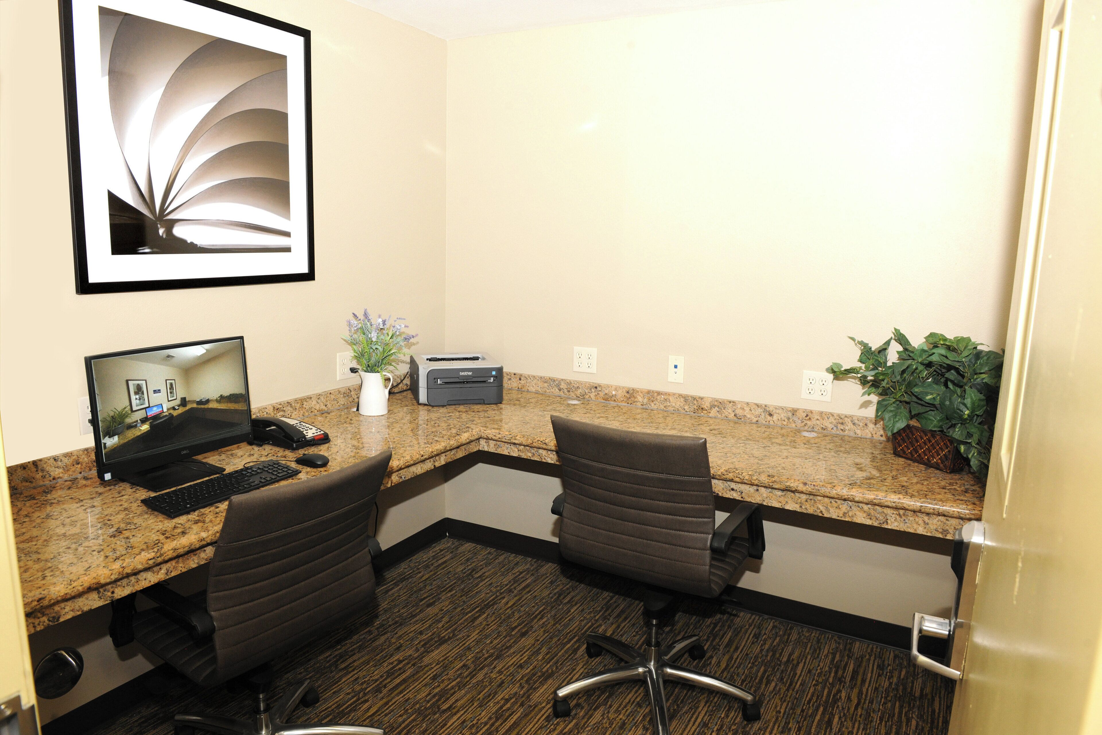 A hotel office with a desktop computer, two chairs, a printer, and a framed picture on the wall.