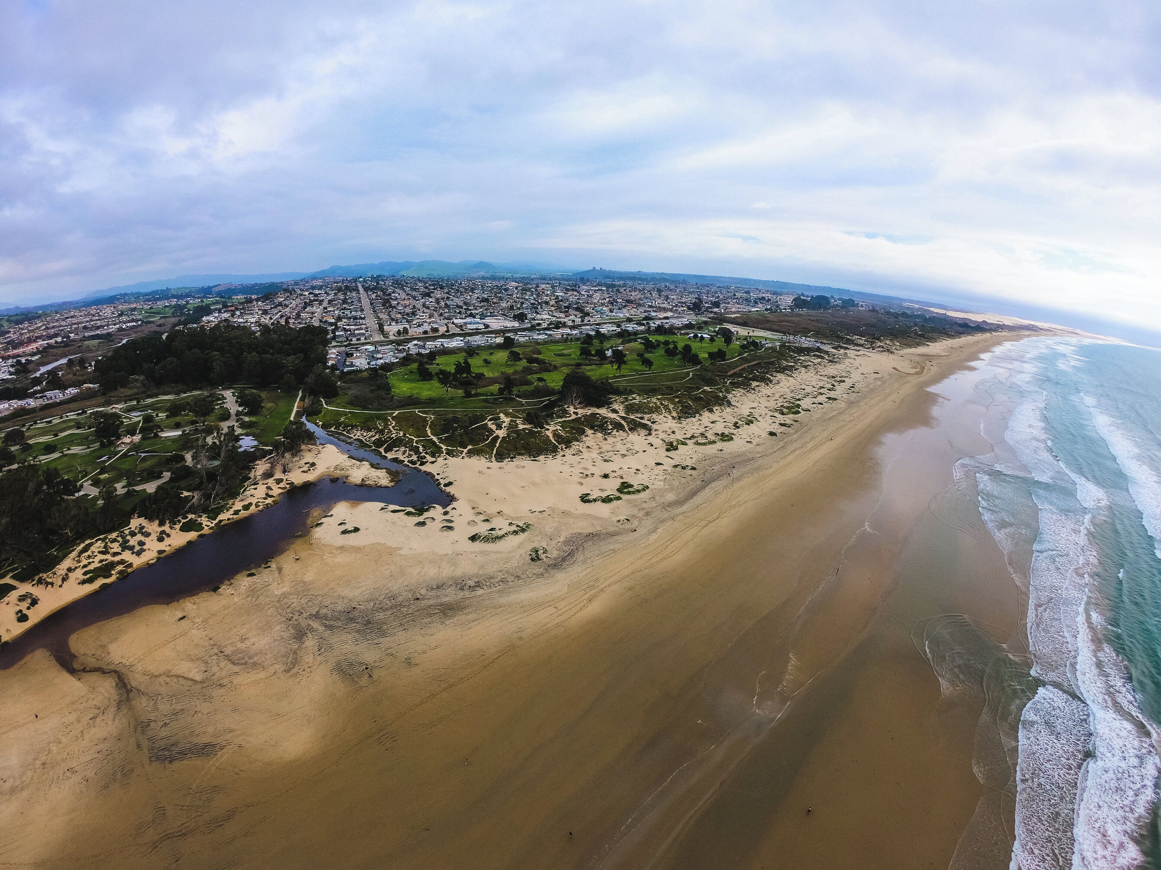 An aerial view of a coastal area with a beach, a park, and a cityscape in the background.