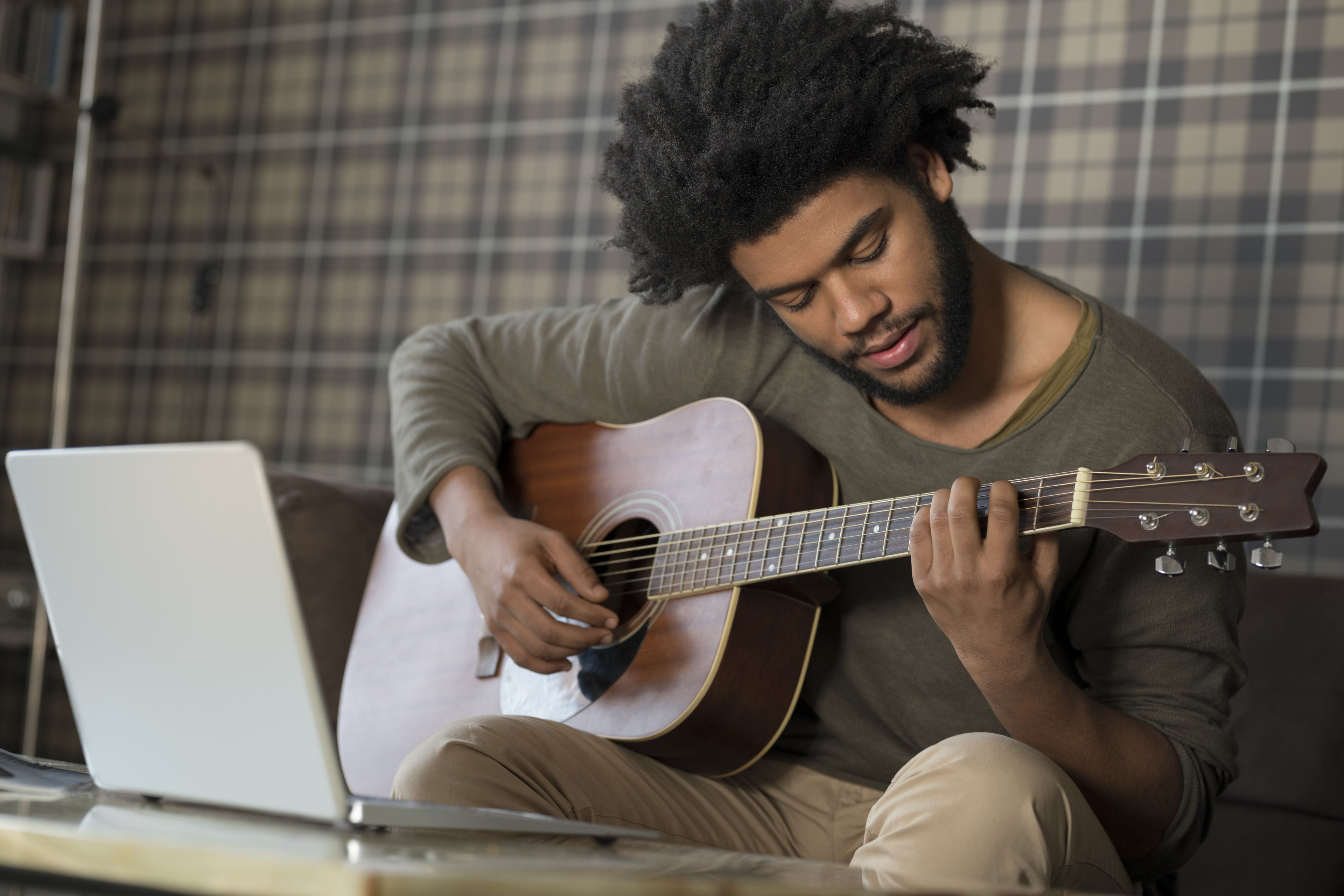 A person sitting on a couch playing a guitar with a laptop on a table in front of them.