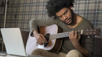 A person sitting on a couch playing a guitar with a laptop on a table in front of them.