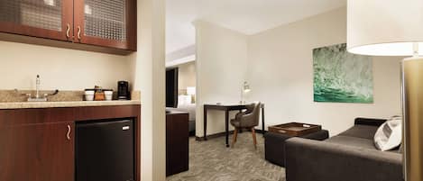 A compact hotel room with a kitchenette, a dining area, and a seating area with a sofa and a lamp.