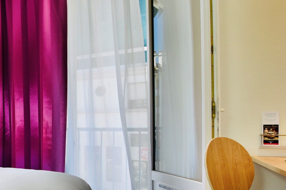 Una habitación de hotel con una cama, una silla de madera, una ventana con cortinas translúcidas y un estante en la pared con un menú.