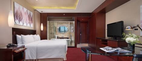 Habitación de hotel con cama, escritorio con computadora, televisión y baño visible a través de una puerta de vidrio.