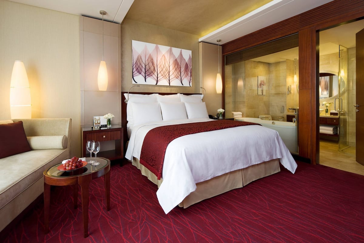 A hotel room with a large bed, a sofa, a small table with a fruit bowl and glasses, and a bathroom visible through a glass door.