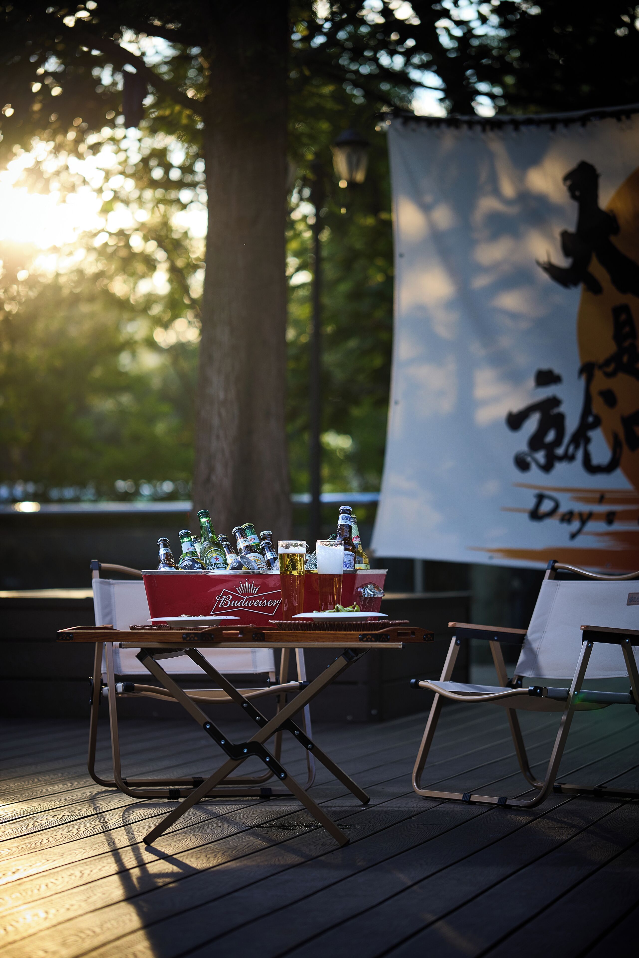 A table with a Budweiser cooler and bottles, two folding chairs, and a banner with Chinese characters in the background.