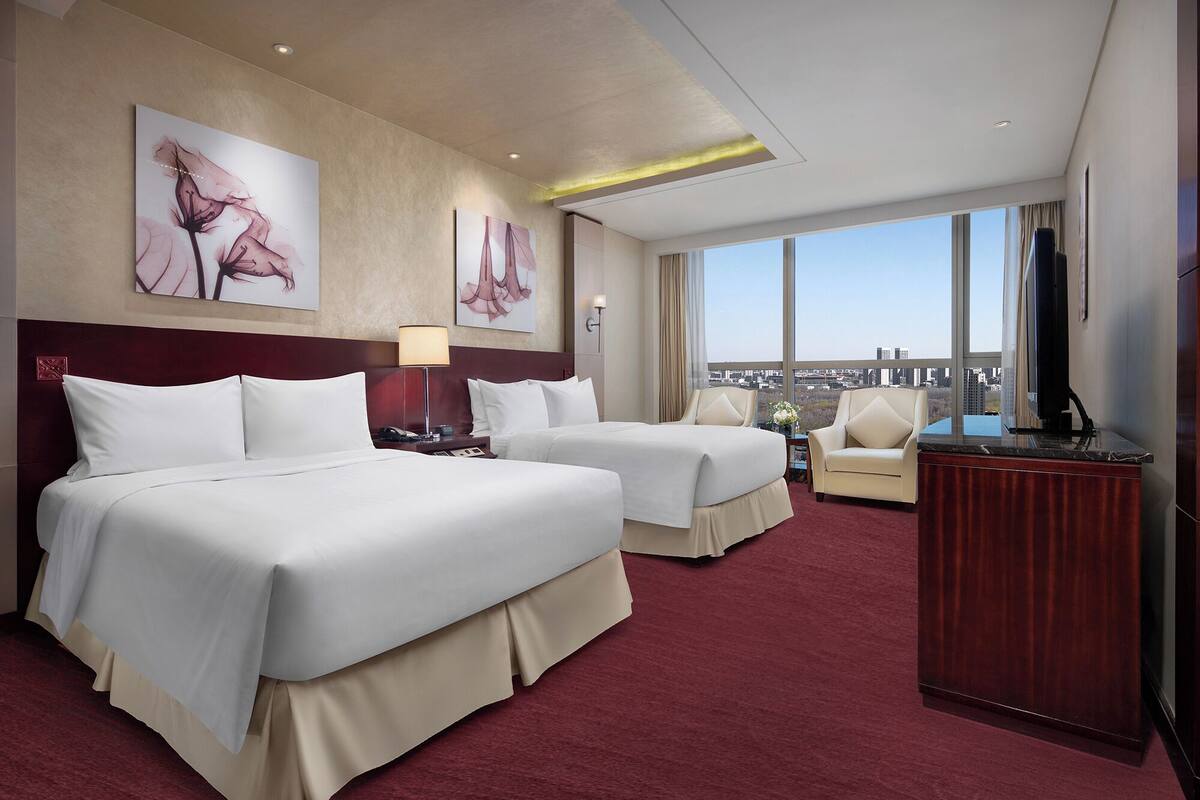 A hotel room with two beds, a desk, a TV, and a view of the city.
