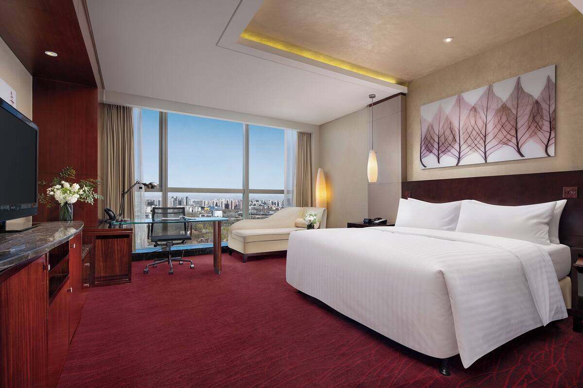 A hotel room with a large bed, a desk with a chair, a TV, a desk lamp, a vase with flowers, and a city view.