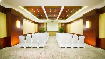 A conference room with rows of white chairs, a large screen at the front, and wooden paneling on the walls.