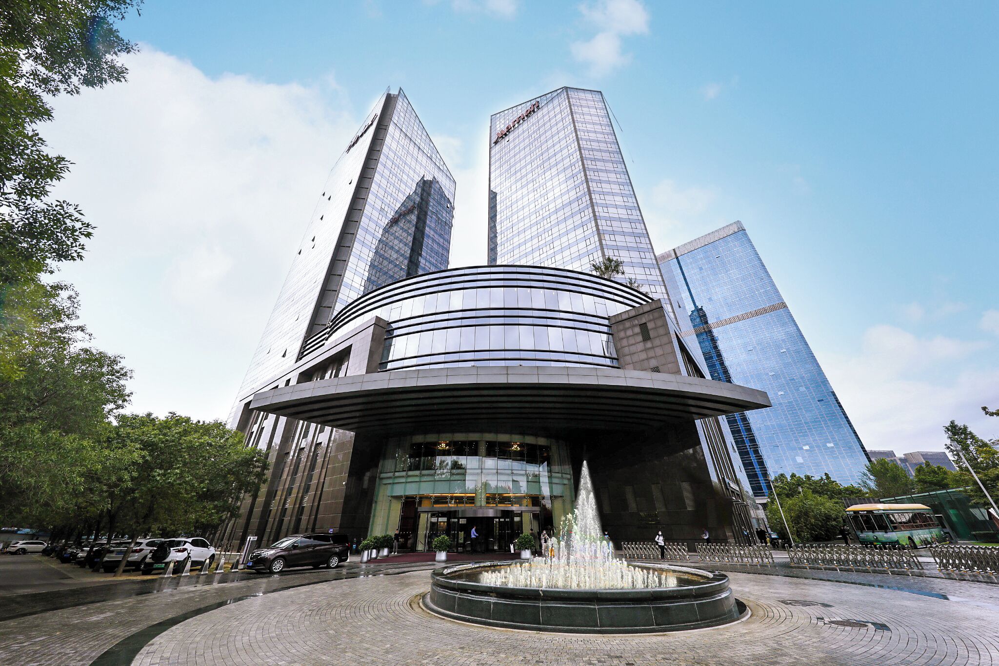 Un edificio moderno con una fuente circular en primer plano, rodeado de árboles y coches estacionados.