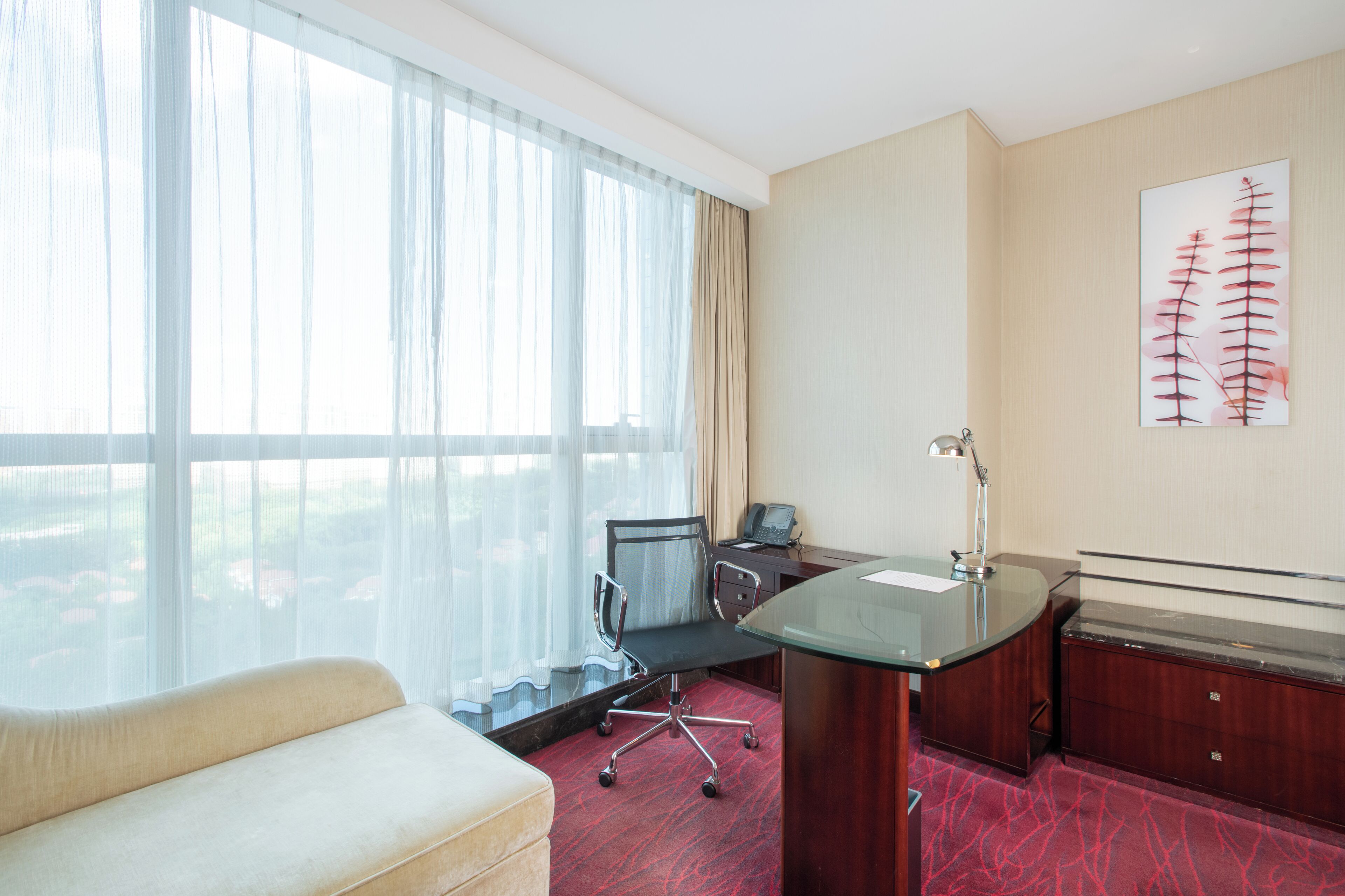 A hotel room with a large window, a desk with a chair, a sofa, and a painting on the wall.
