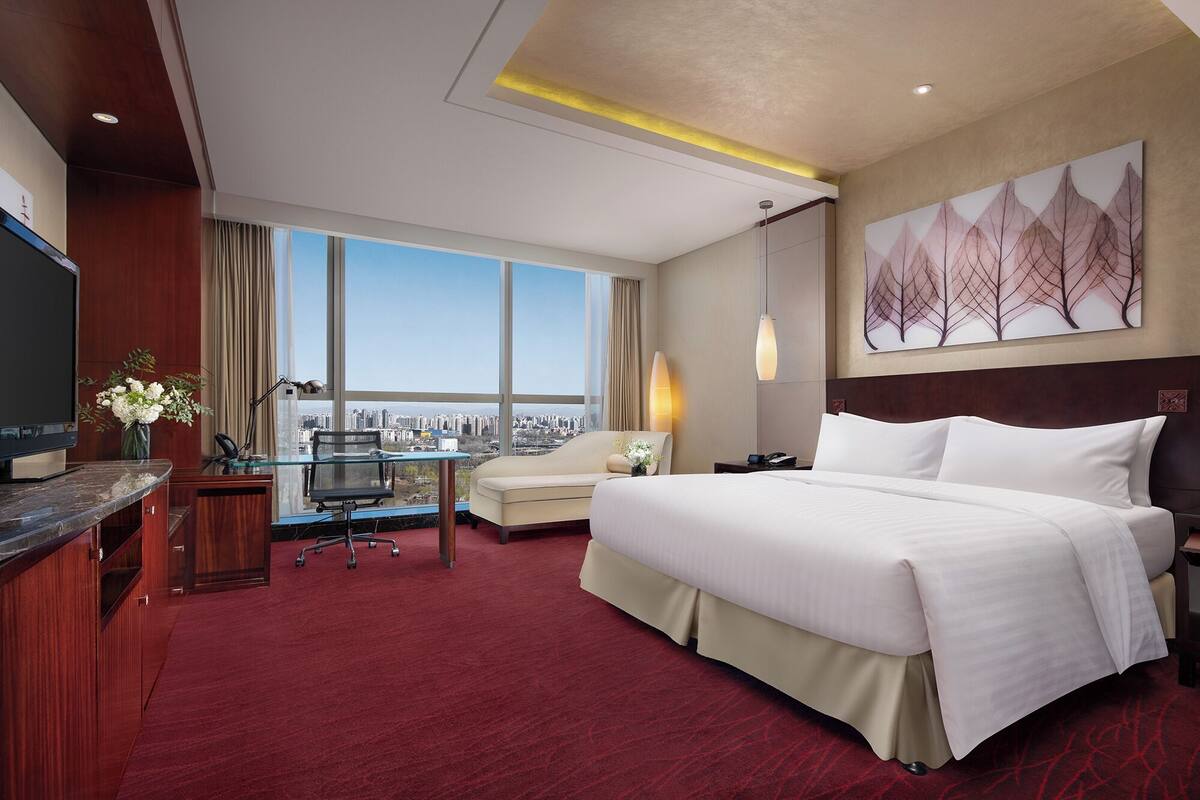 A hotel room with a bed, desk, chair, and a large window with a city view.