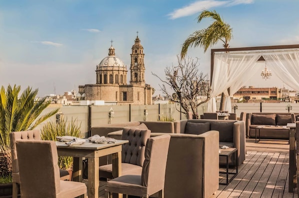 Zona de estar al aire libre con mesas y sillas, un toldo blanco y vista de un edificio histórico con cúpulas y torres.