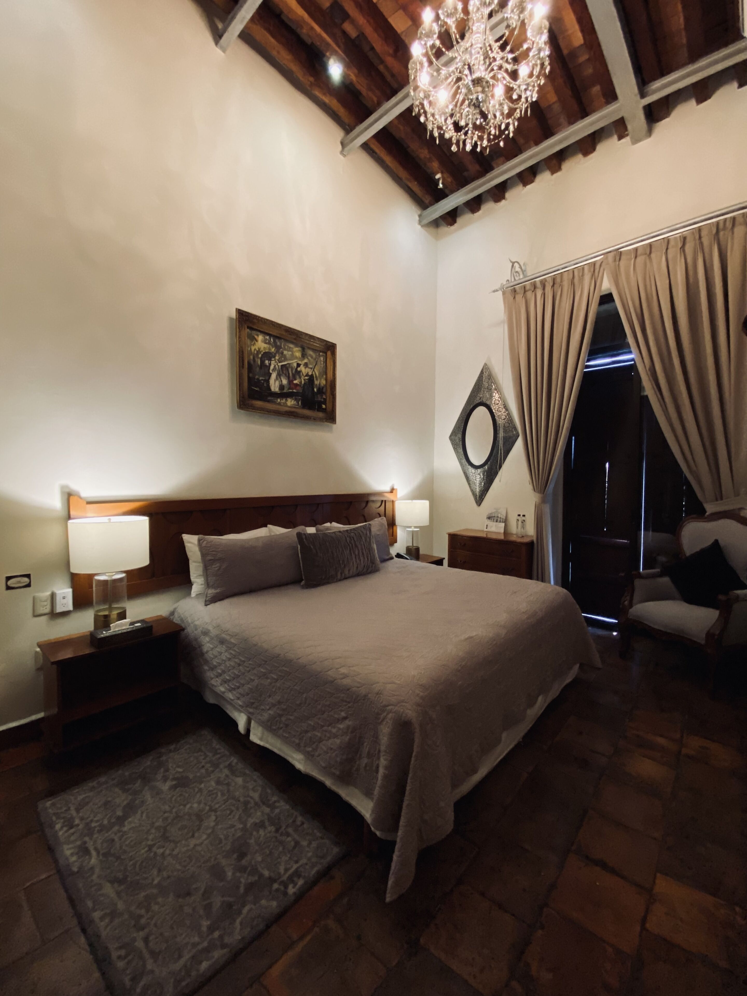 A bedroom with a large bed, a chandelier, a painting on the wall, and a view through the curtains.