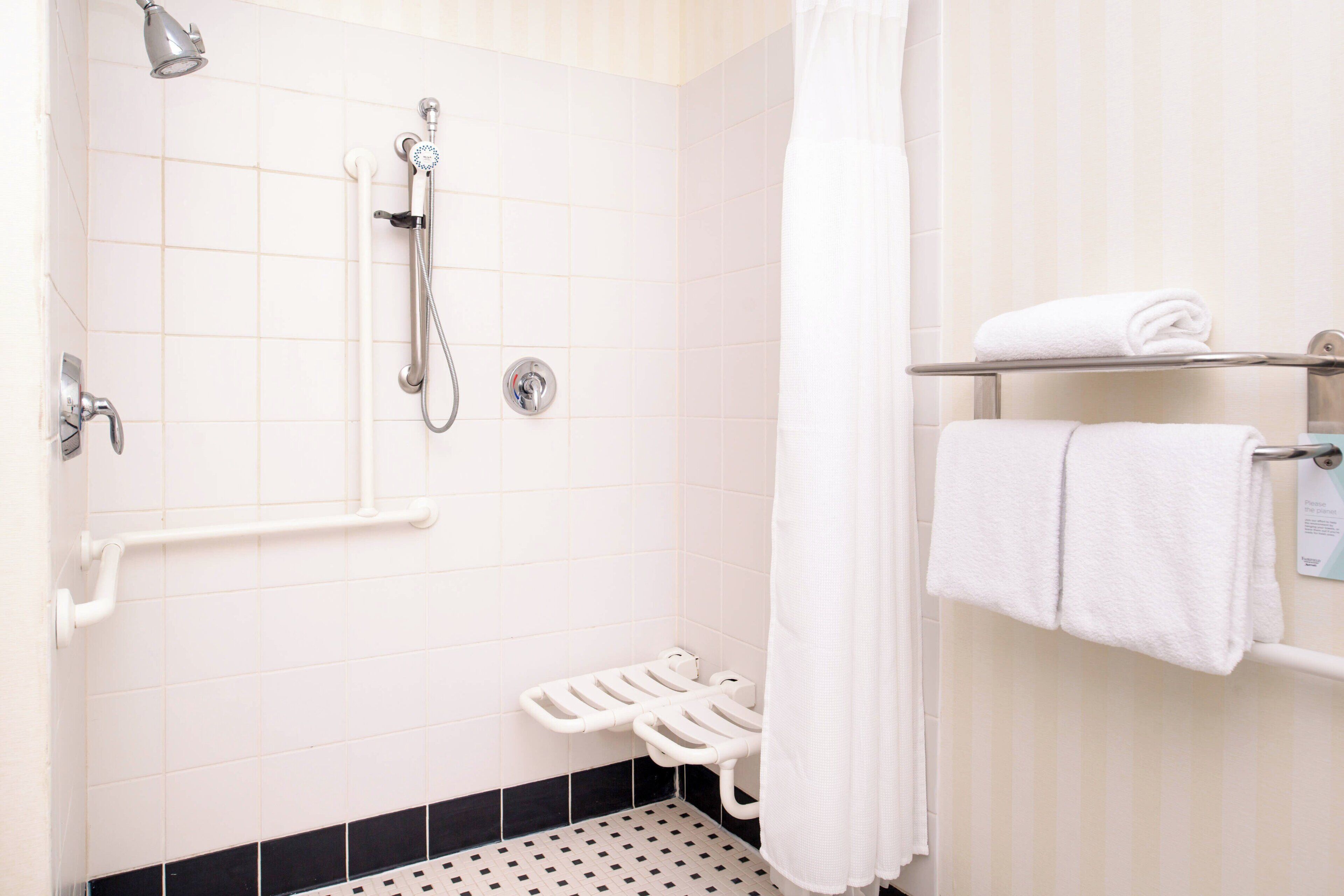 A bathroom with white tiles, a shower with a handheld showerhead, a toilet with a grab bar, and a towel rack with folded towels.