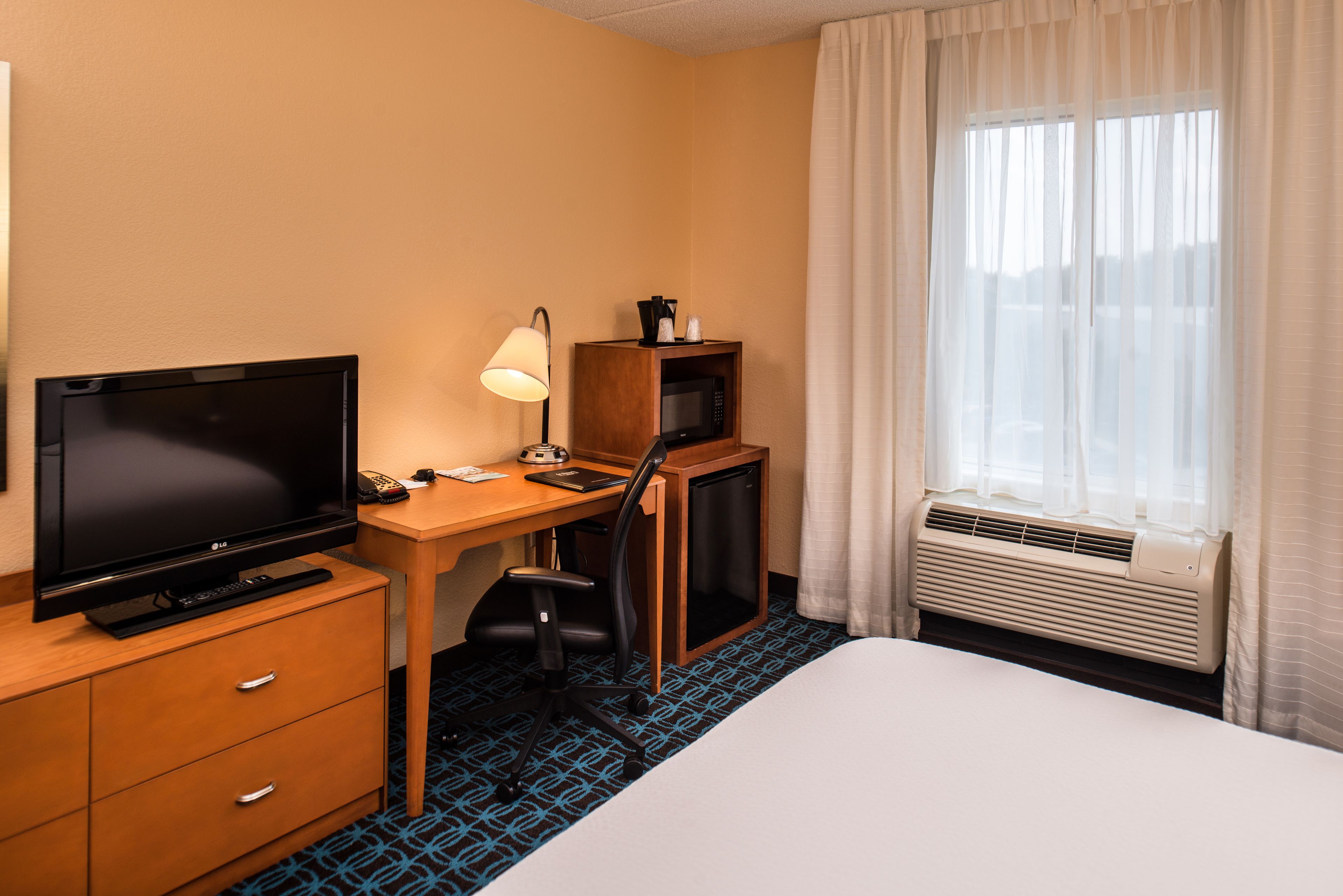Une chambre d’hôtel équipée d’un lit, d’un bureau avec un ordinateur, d’une télévision, d’une lampe, d’un four à micro-ondes et d’un appareil de climatisation.