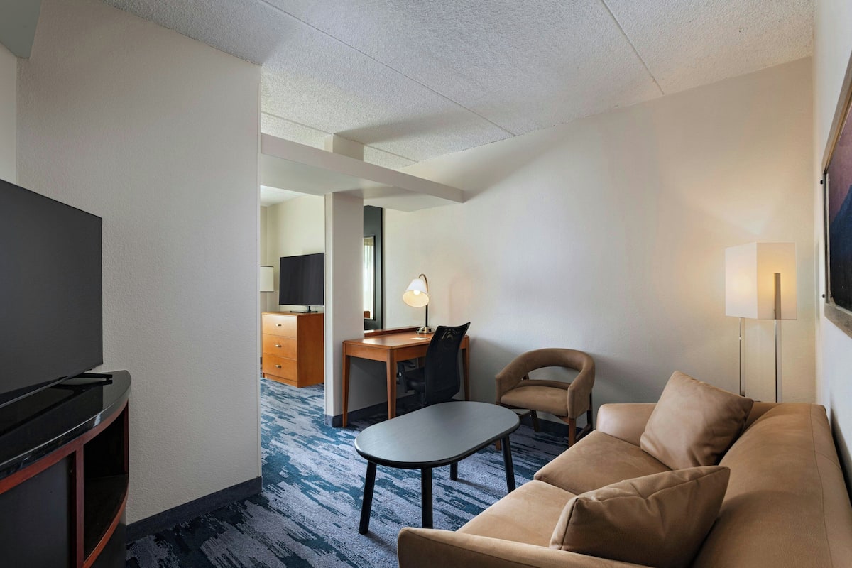 Ein Hotelzimmer mit Flachbildfernseher, einer Sitzecke, einem kleinen Tisch, einem Schreibtisch mit Stuhl und einer Lampe.