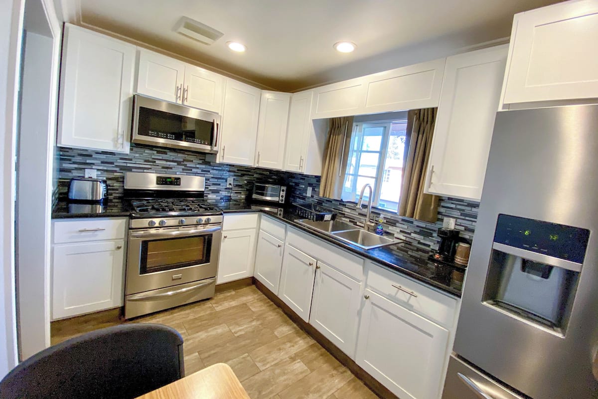 A modern kitchen with white cabinets, stainless steel appliances, a tiled backsplash, and a window with curtains.