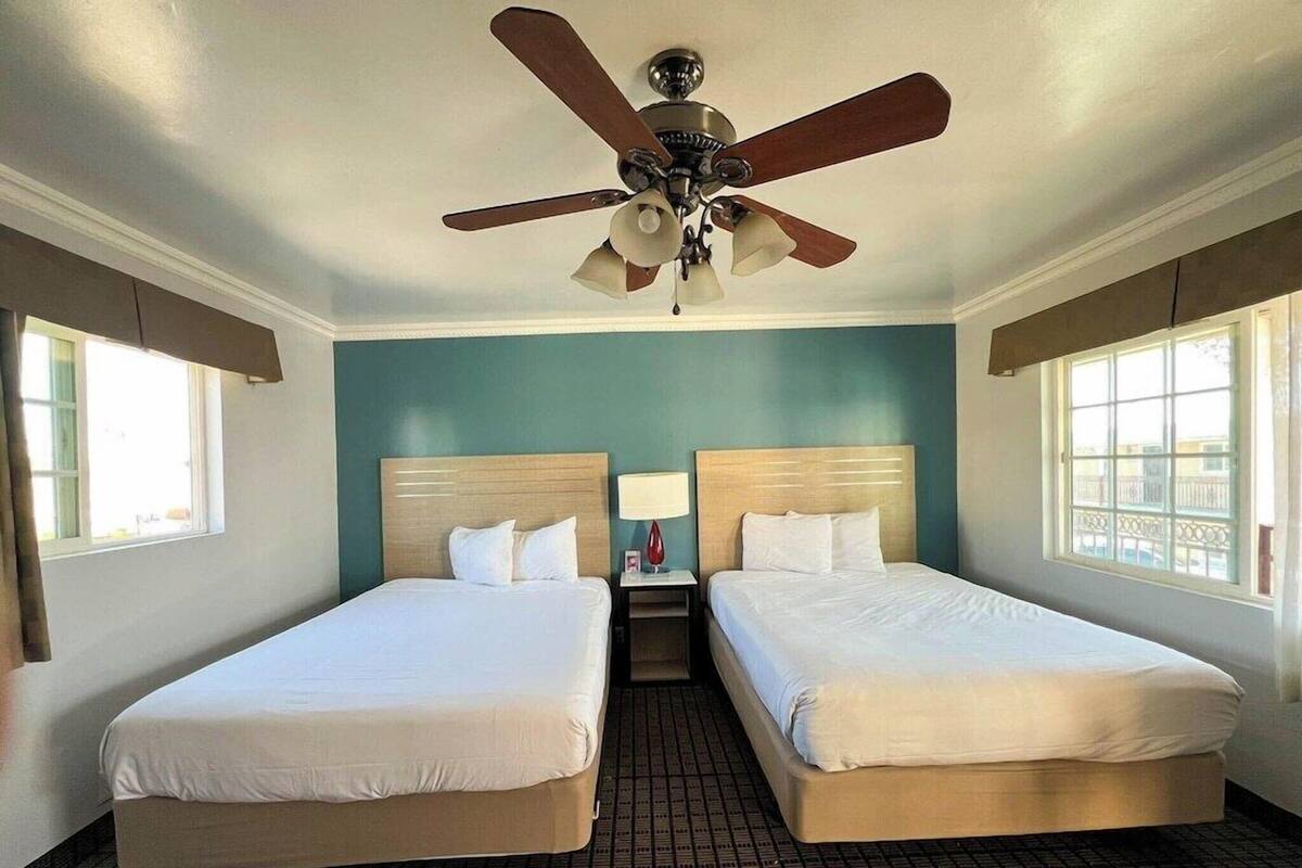 A hotel room with two beds, a ceiling fan, a nightstand with a lamp, and a window with a view.