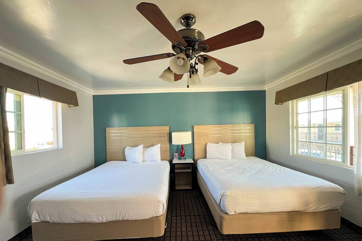 A hotel room with two single beds, a ceiling fan, a nightstand with a lamp, and a window with a view.