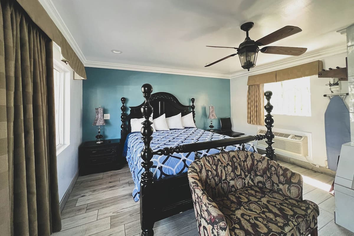 A bedroom with a four-poster bed, a ceiling fan, a patterned armchair, and a window with curtains.