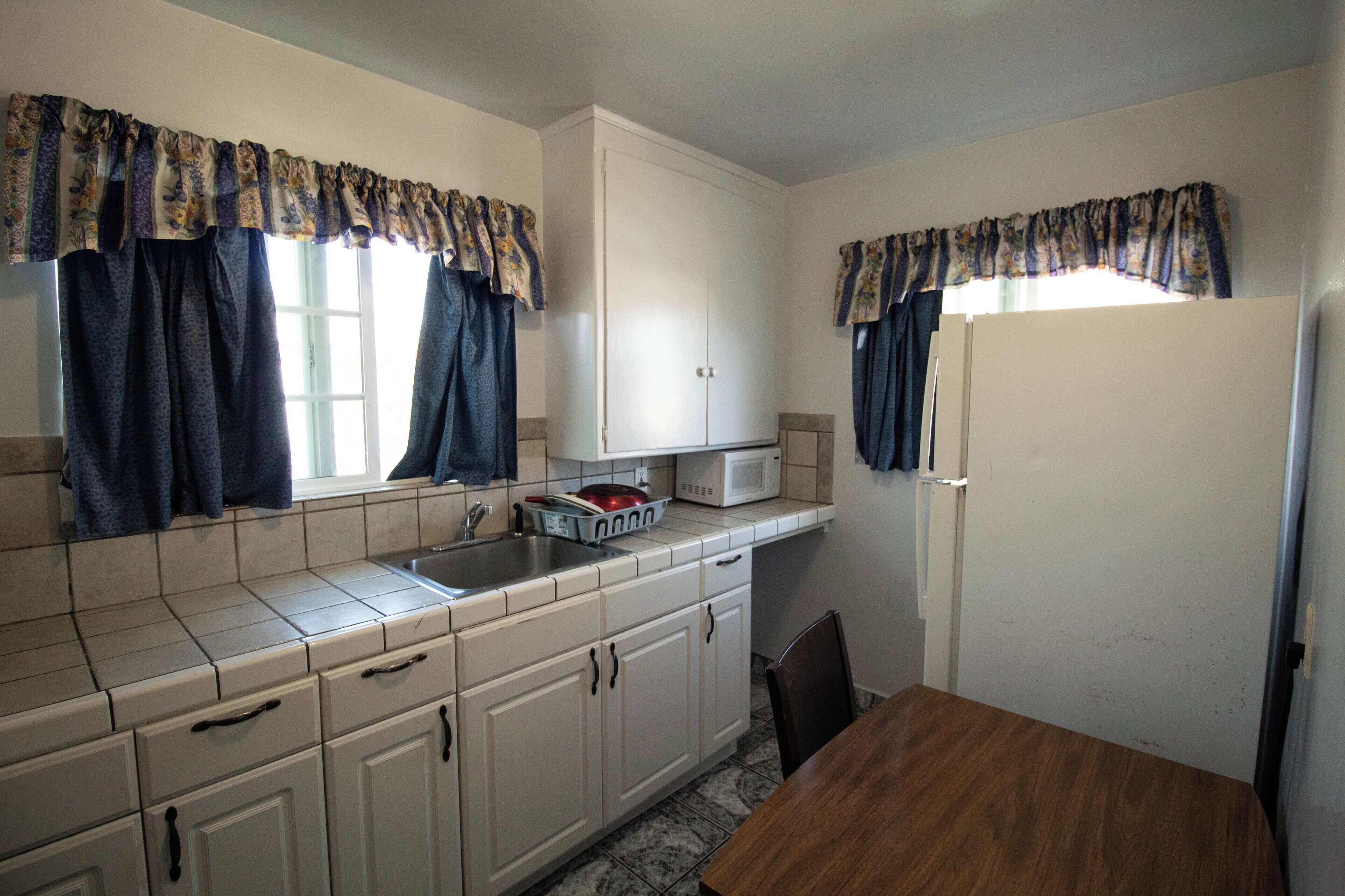 A compact kitchen with white cabinets, a microwave, and a refrigerator. There is a window with curtains, a sink, and a dining table with chairs.