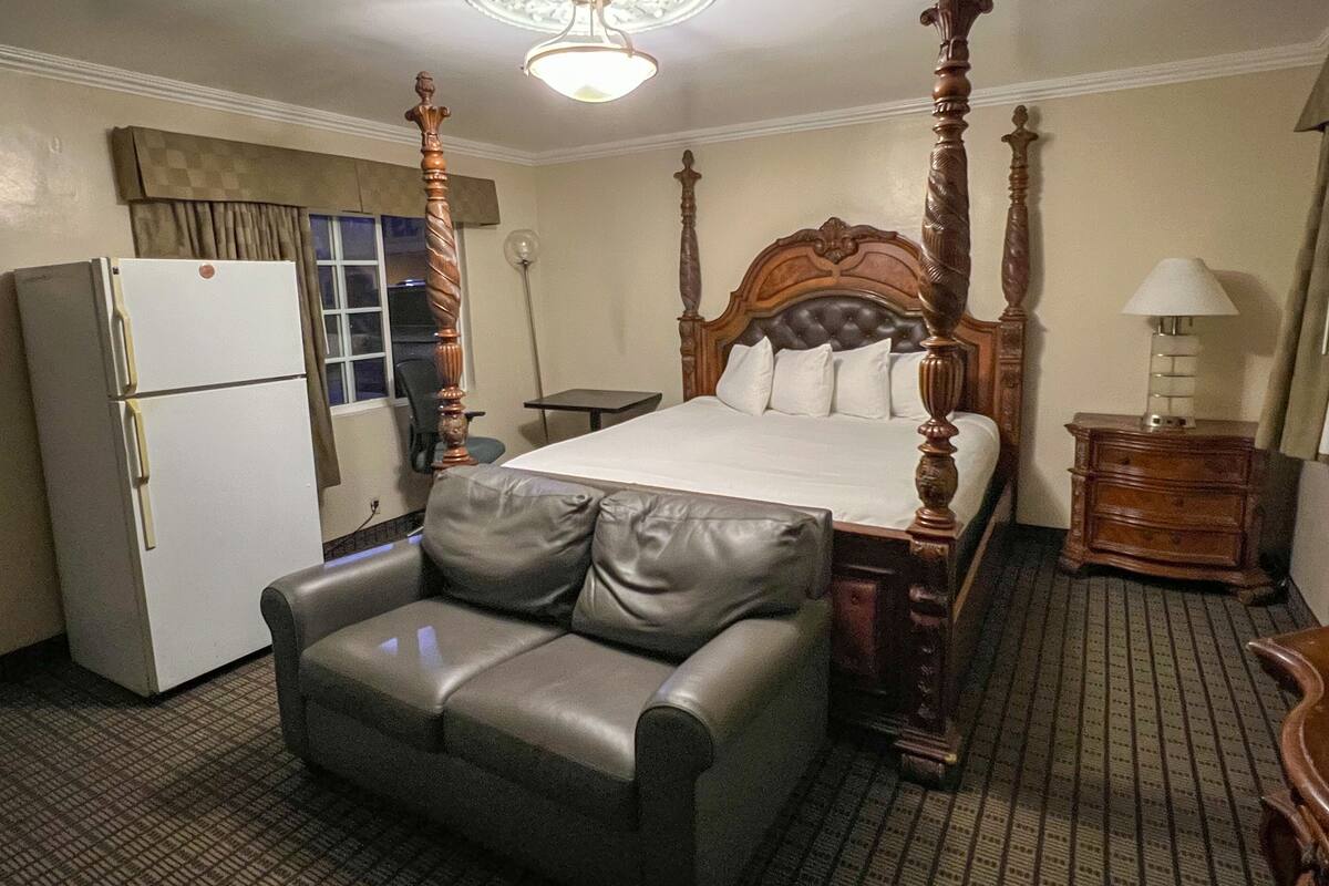 A hotel room with a four-poster bed, a refrigerator, a chair, a bedside table with a lamp, and a ceiling light fixture.