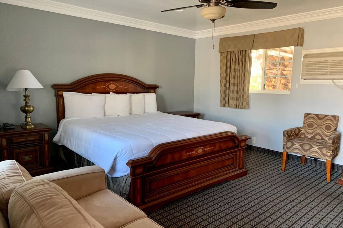 A hotel room with a wooden bed, a nightstand with a lamp, a patterned armchair, and a window with curtains.