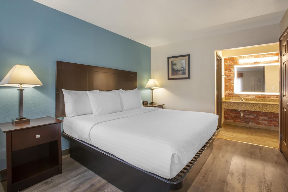 A hotel room with a large bed, two bedside tables with lamps, a small nightstand, a bathroom with a mirror and sink, and a brick accent wall.