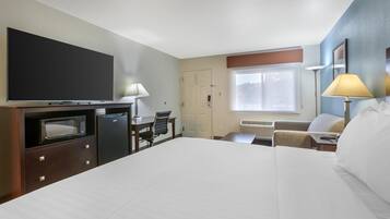 A hotel room with a large bed, a desk with a chair, a microwave, a refrigerator, a flat-screen TV, and a window with blinds.