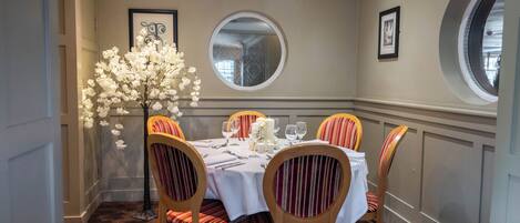 A dining area with a round table, chairs, a vase of flowers, framed artwork, and a round mirror on the wall.