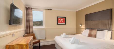 A hotel room with a bed, a wooden dresser, a chair, a television mounted on the wall, and a window with curtains.