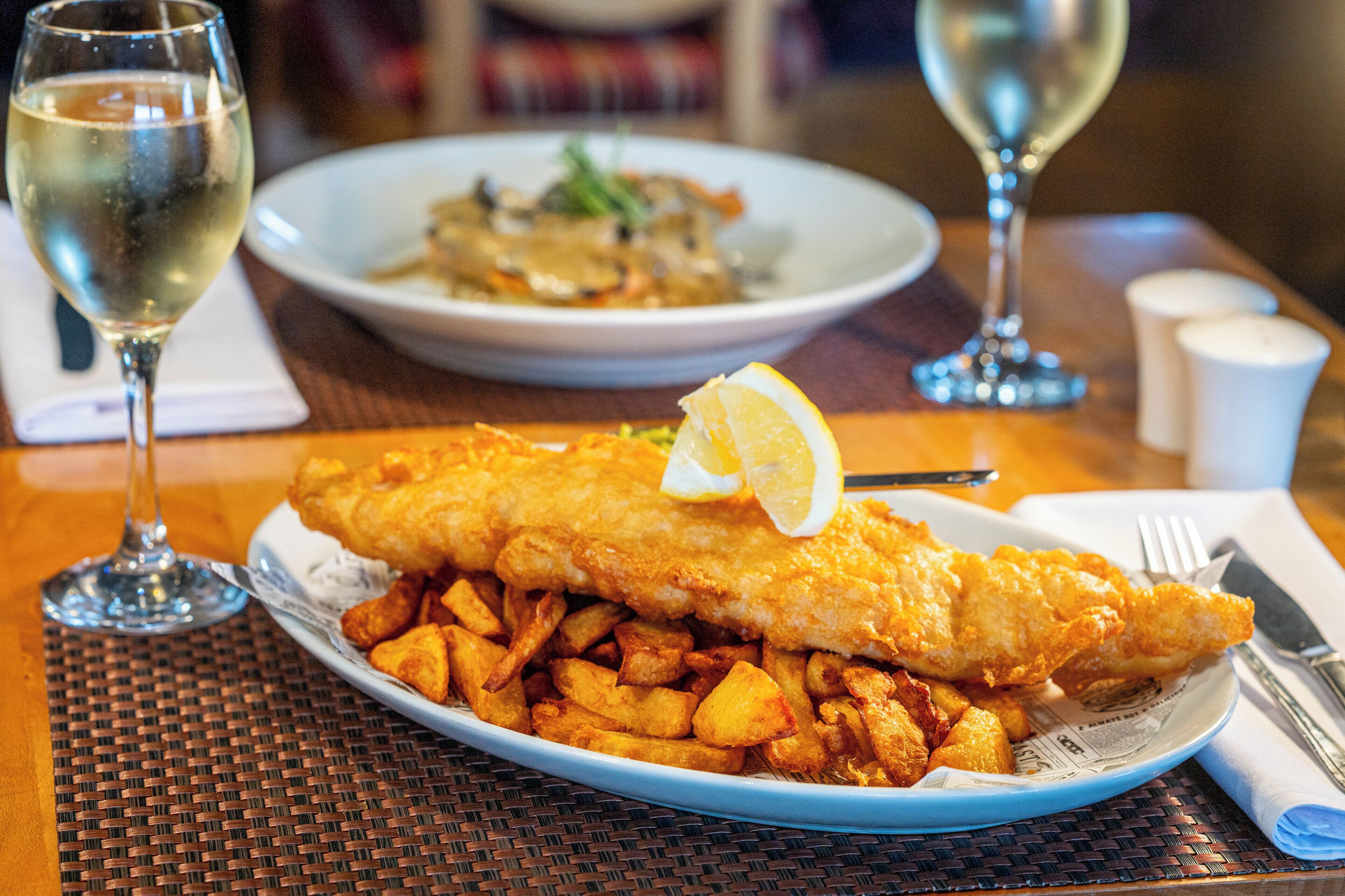 A plate of fish and chips with a lemon wedge, accompanied by a glass of white wine, on a table with a woven placemat.