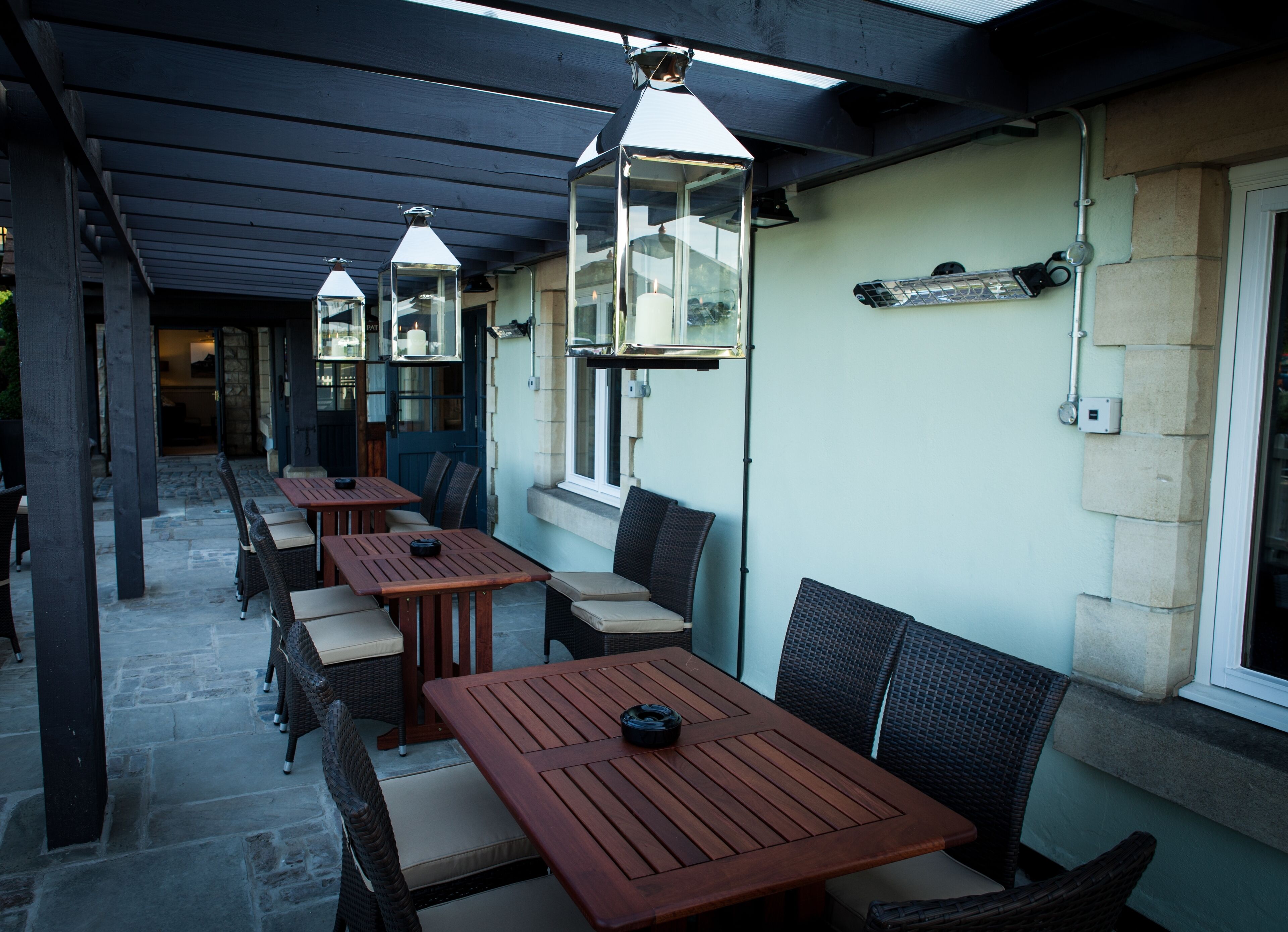 An outdoor patio area with wooden tables and wicker chairs, under a covered roof with hanging lanterns.