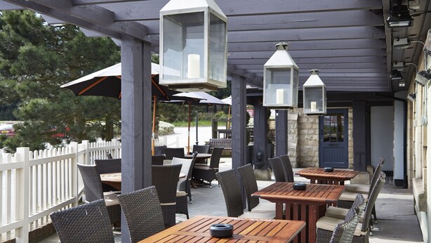 An outdoor patio with wooden tables and wicker chairs, white lanterns hanging above, and a white picket fence in the background. - Red Lion Todwick by Chef and Brewer Collection (Sheffield)