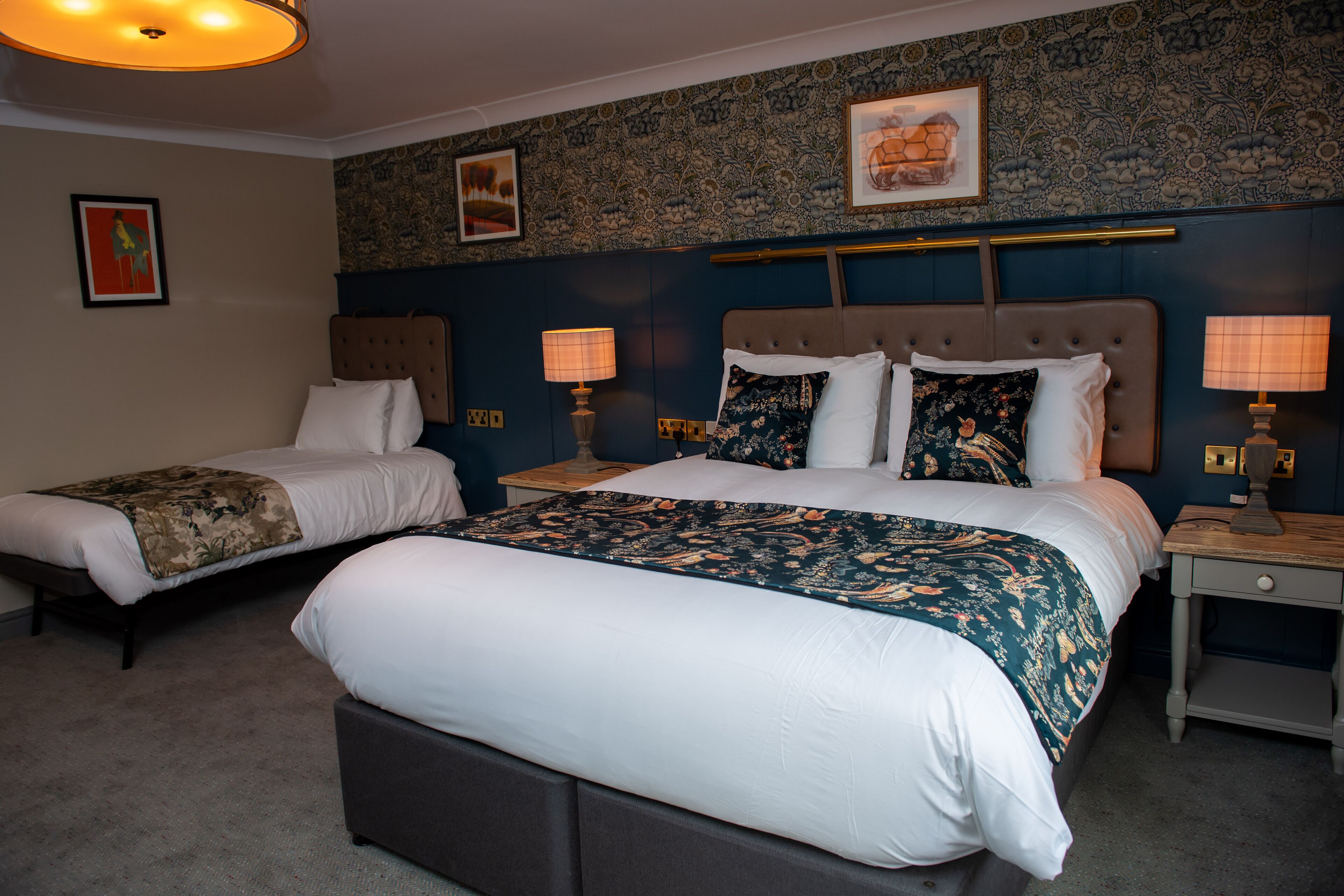 A hotel room with two beds, a large bed with a floral bedspread and a smaller bed with a solid-colored bedspread. There are two bedside tables with lamps, a headboard, and a wall with floral wallpaper.