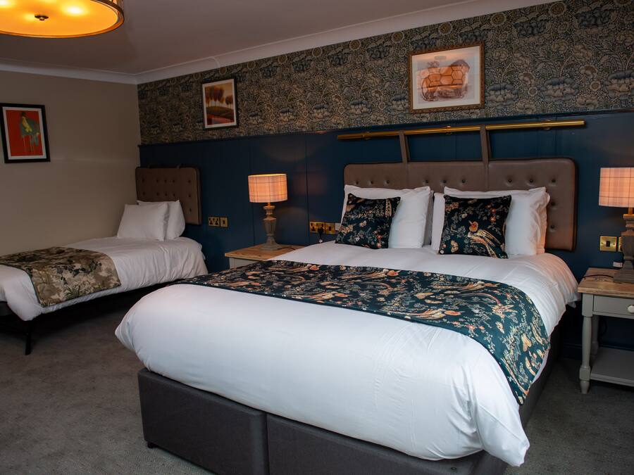 A hotel room with two beds, a large bed with a floral bedspread and a smaller bed with a solid-colored bedspread. There are two bedside tables with lamps, a headboard, and a wall with floral wallpaper.