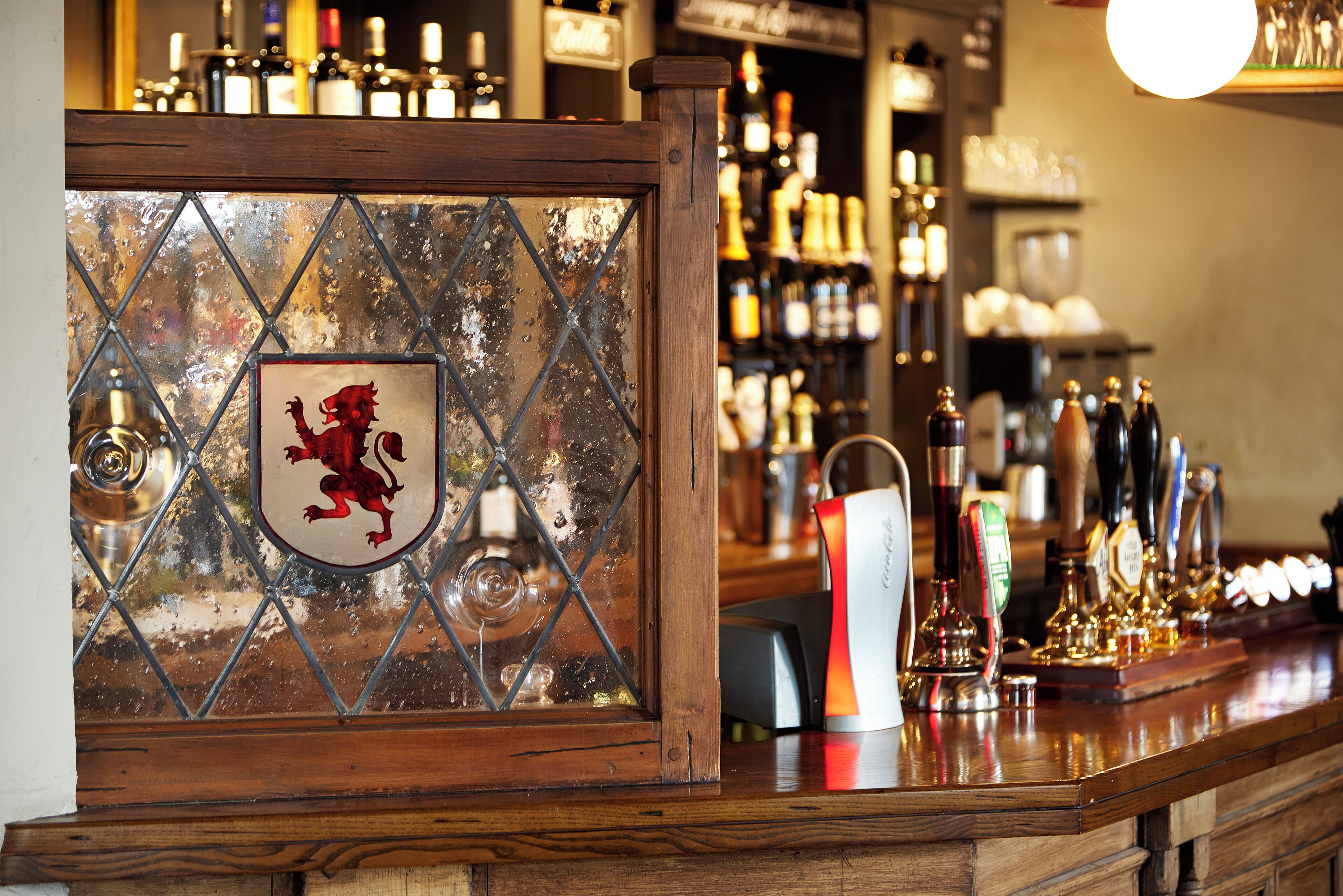 A bar area with a wooden counter, a shield with a red dragon emblem, and a variety of bottles and taps.