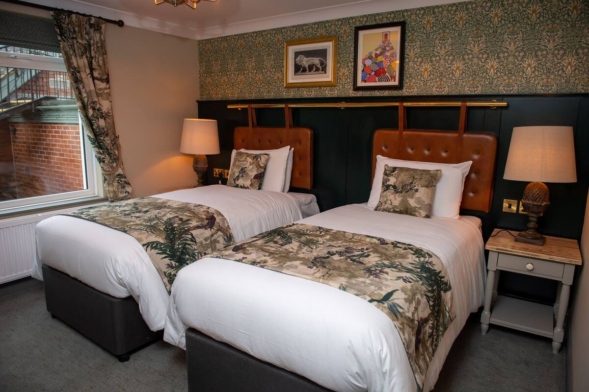 A hotel room with two beds, a window with a view of a brick building, and a patterned wallpaper.