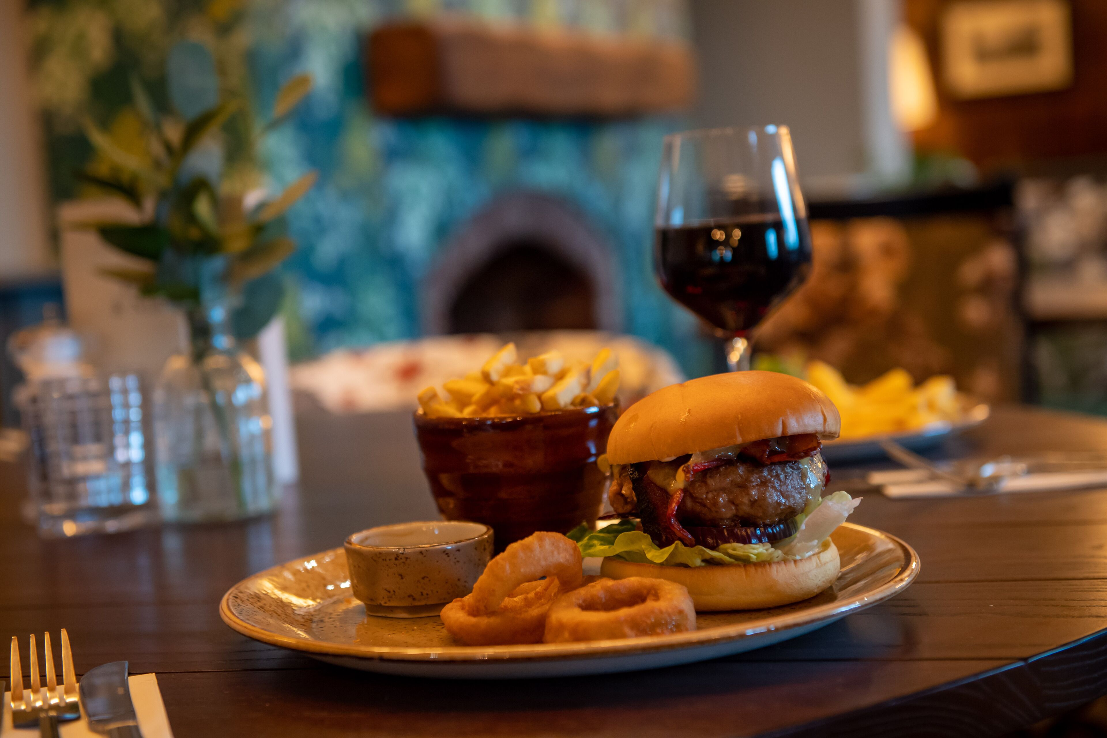 A dining table with a burger, fries, onion rings, and a glass of red wine.