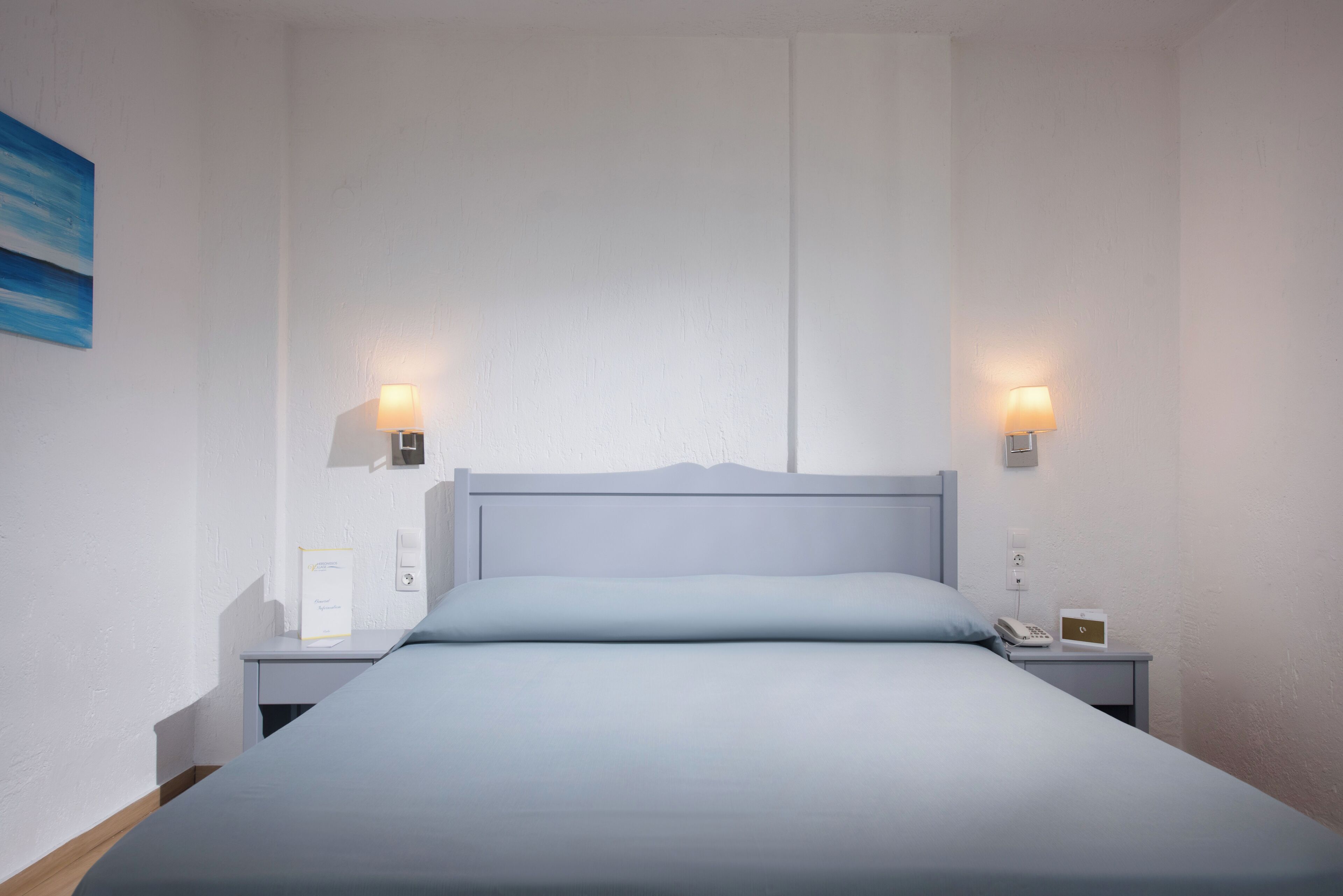 Una cama bien tendida con una colcha azul claro y almohadas, flanqueada por dos mesitas de noche con lámparas y un pequeño reloj.
