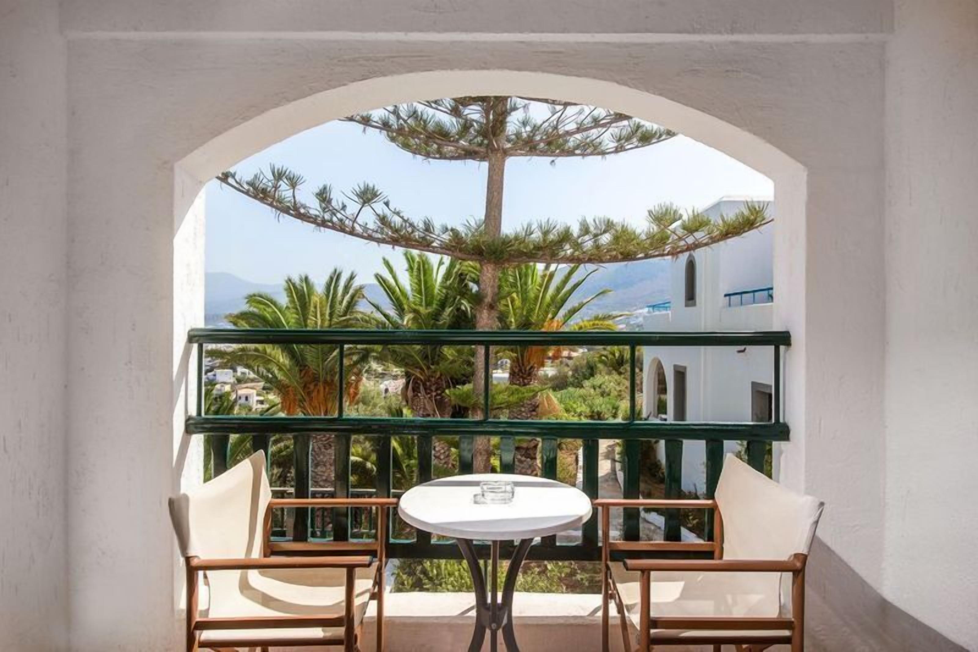 Un balcón con una mesa y dos sillas, con vistas a un jardín con palmeras y un edificio al fondo.