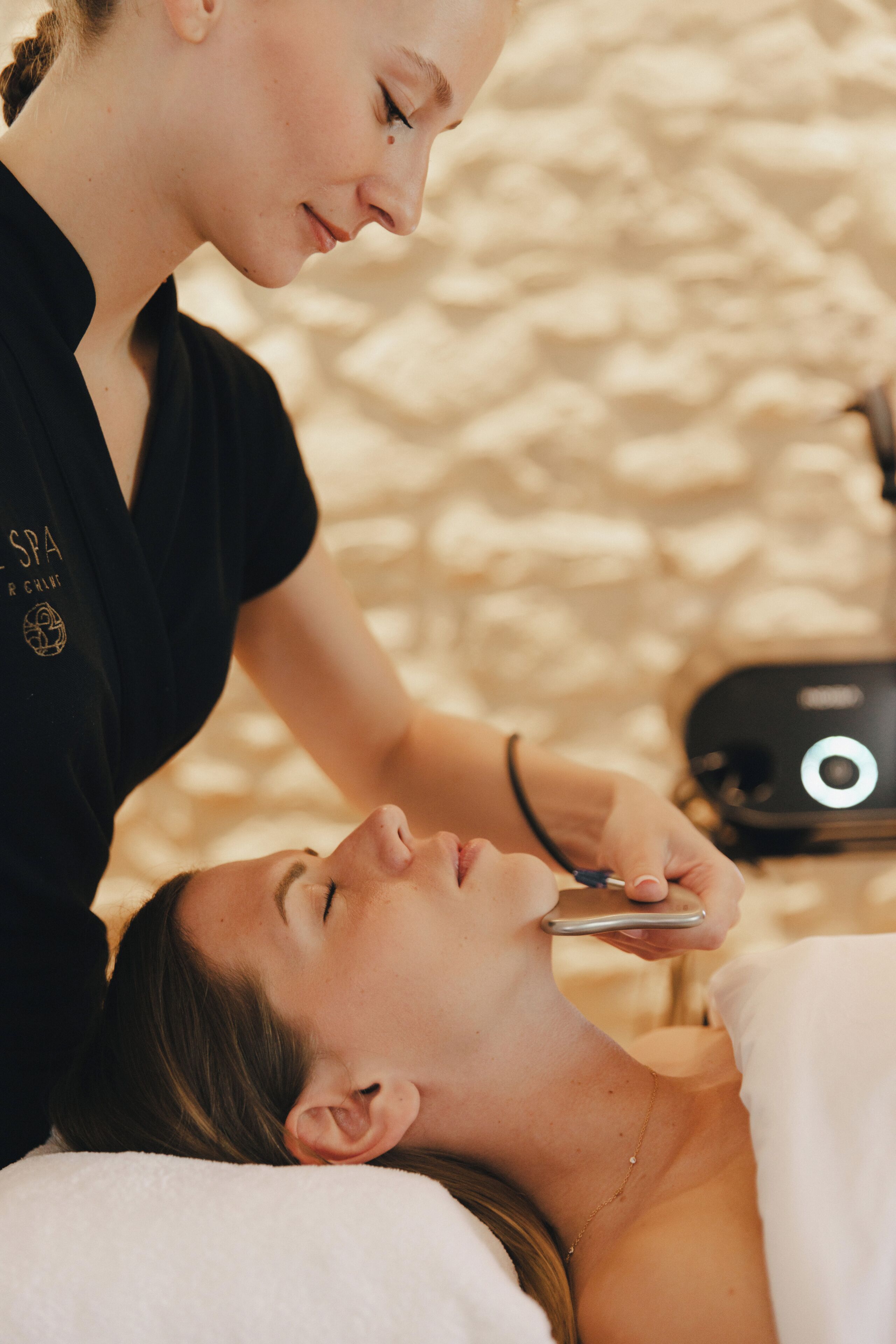 A person lying down with a device being used on their face, another person standing beside them, and a stone wall in the background.