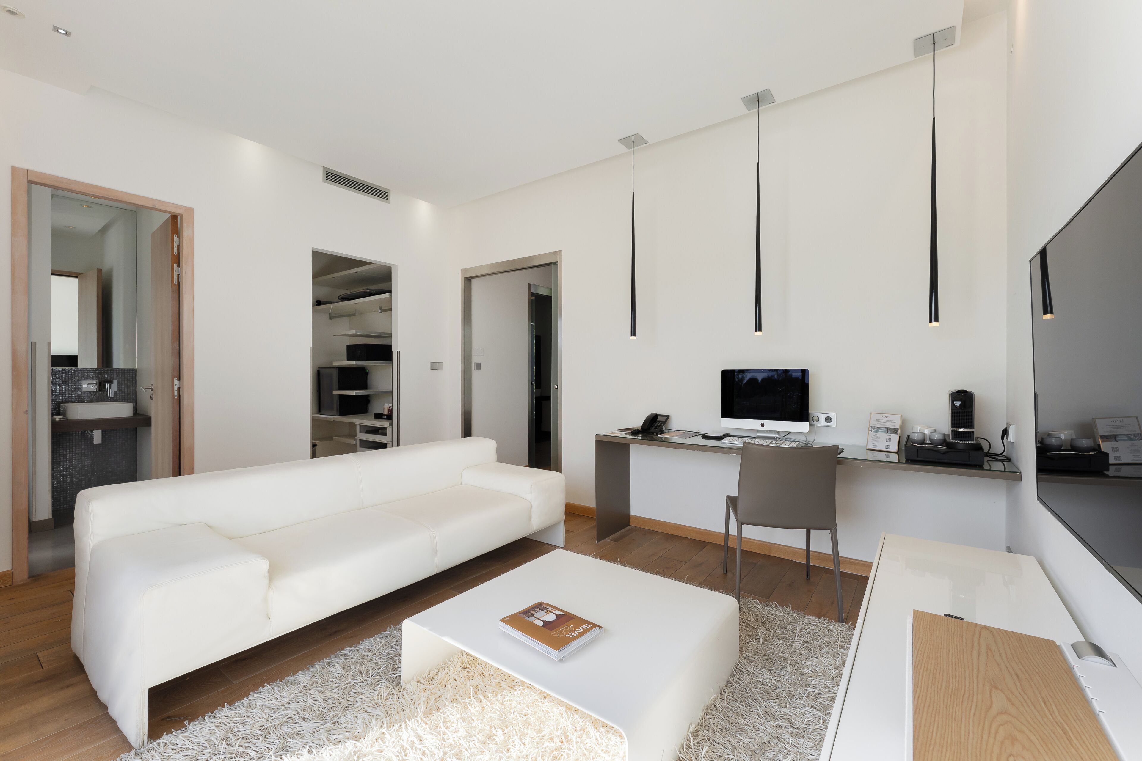 A modern living room with a white sofa, a coffee table, a desk with a computer, and a TV. There is a bookshelf and a kitchen area visible in the background.