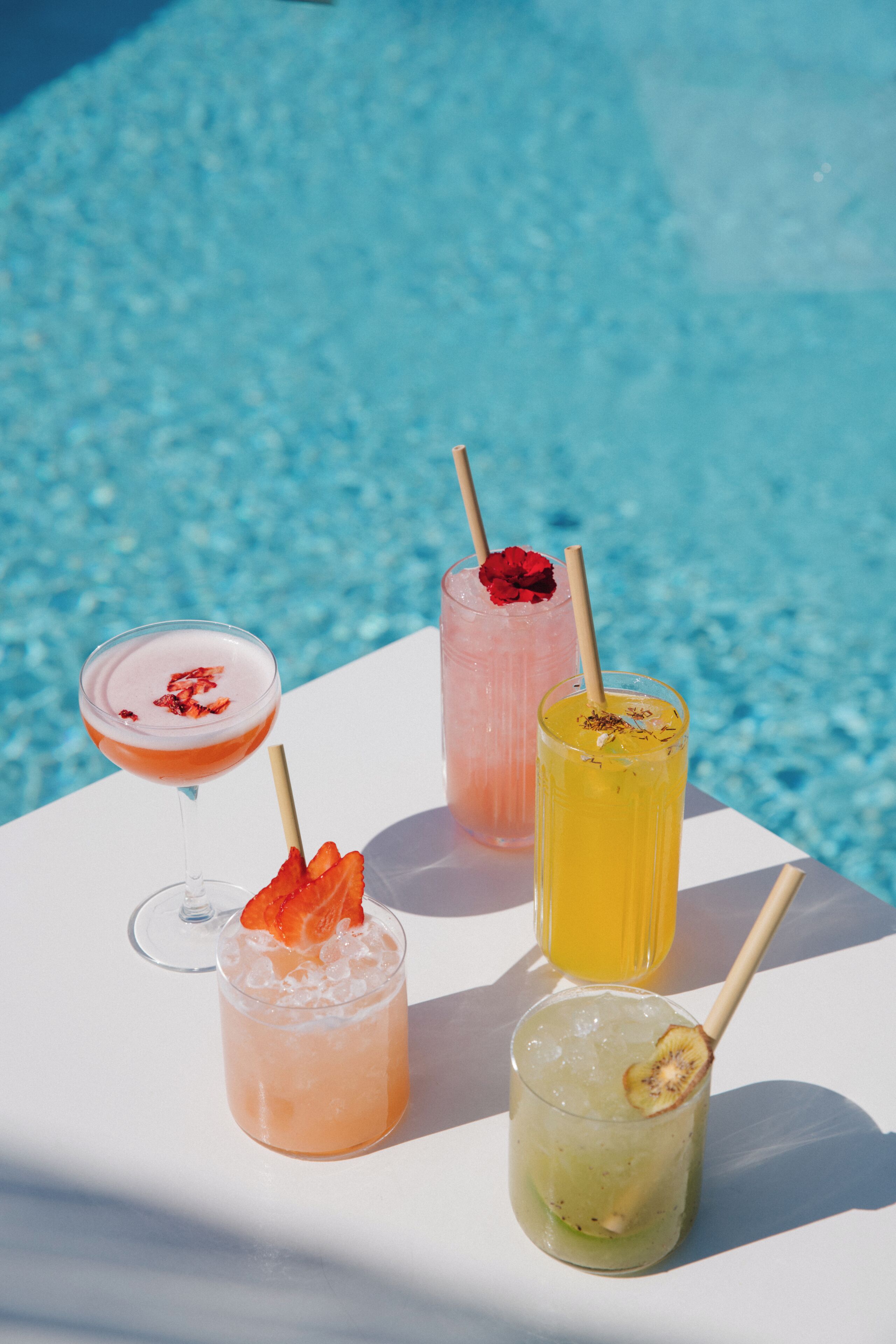 Four cocktails on a white table by a pool.