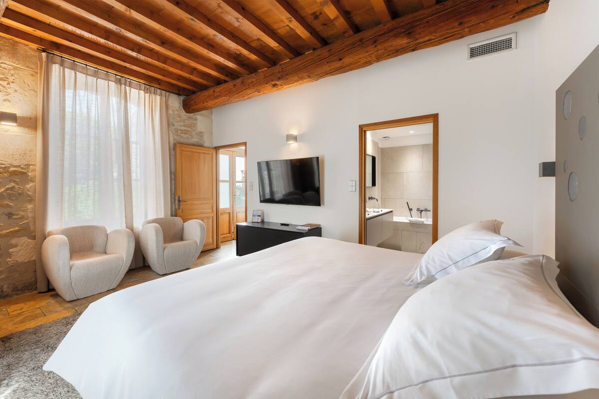 Een slaapkamer met een groot bed, een televisie, twee fauteuils, een stenen muur en een houten plafond.