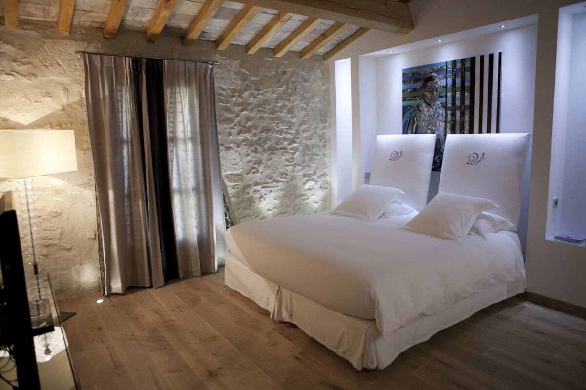 Een slaapkamer met een stenen muur, een bed met witte lakens, een houten vloer en een groot raam met gordijnen.