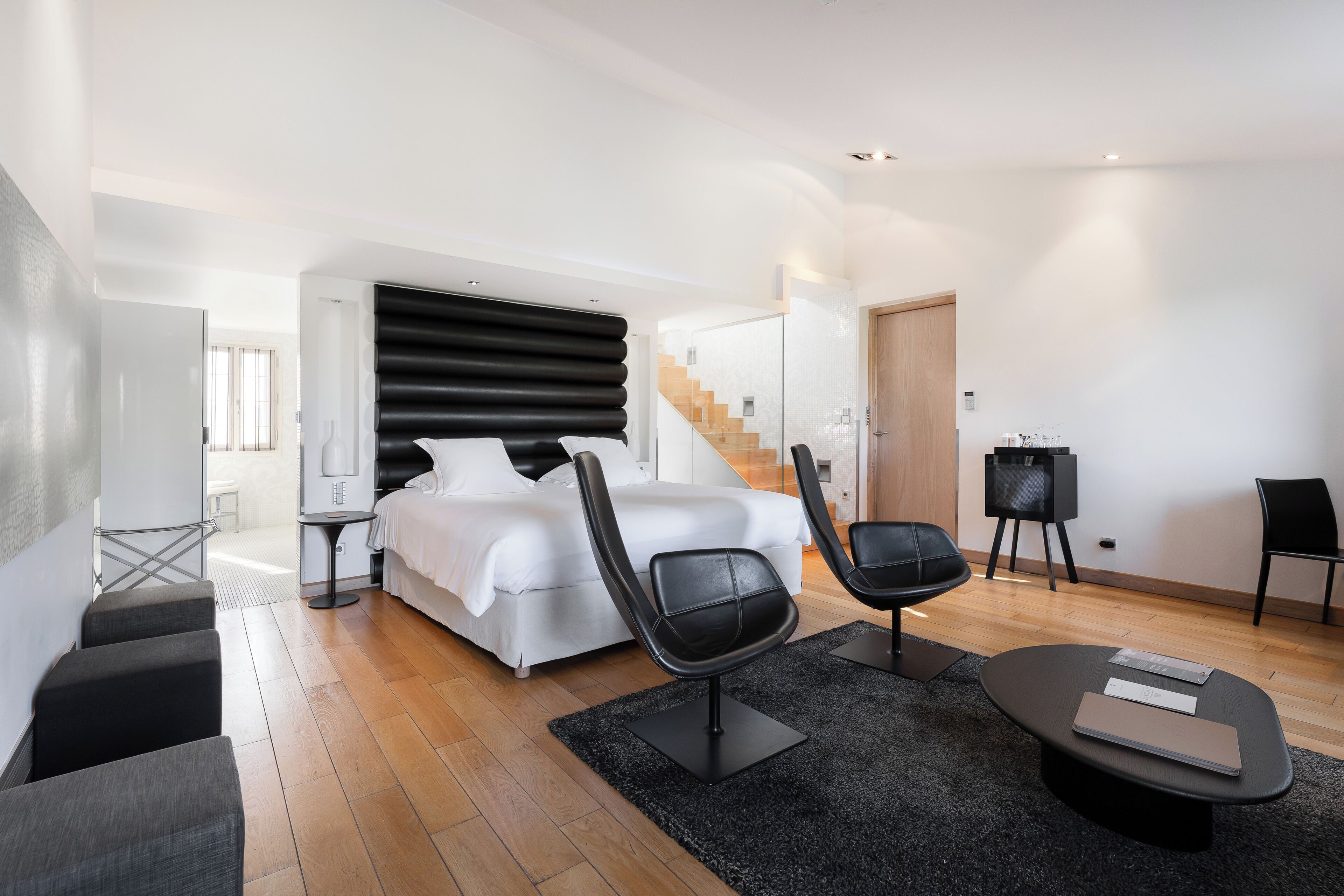 A modern bedroom with a bed, two black chairs, a small table, and a staircase leading to another room.