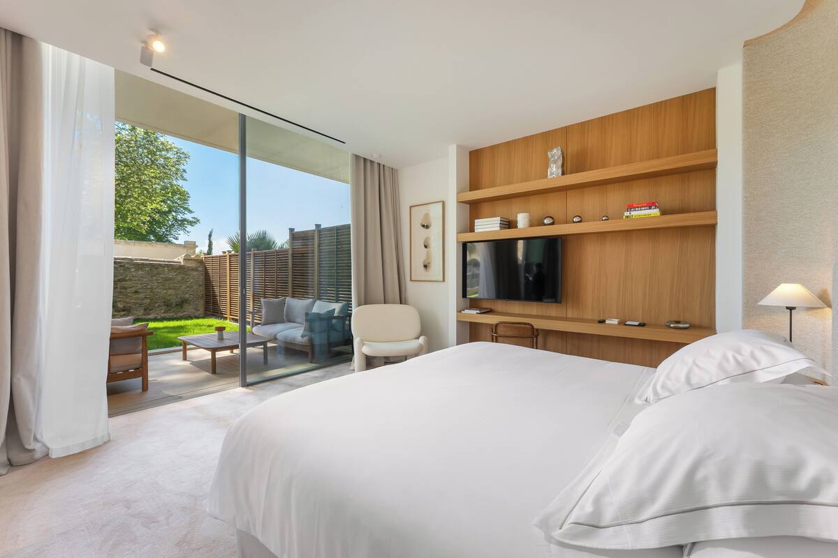 Een moderne hotelkamer met een groot bed, een televisie, een houten plank met spullen, een glazen deur naar een balkon en een klein tafeltje met stoelen.
