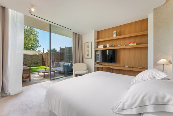 Una habitación de hotel moderna con una cama grande, un televisor, un estante de madera con objetos, un ventanal de vidrio que da a un balcón y una pequeña mesa con sillas.