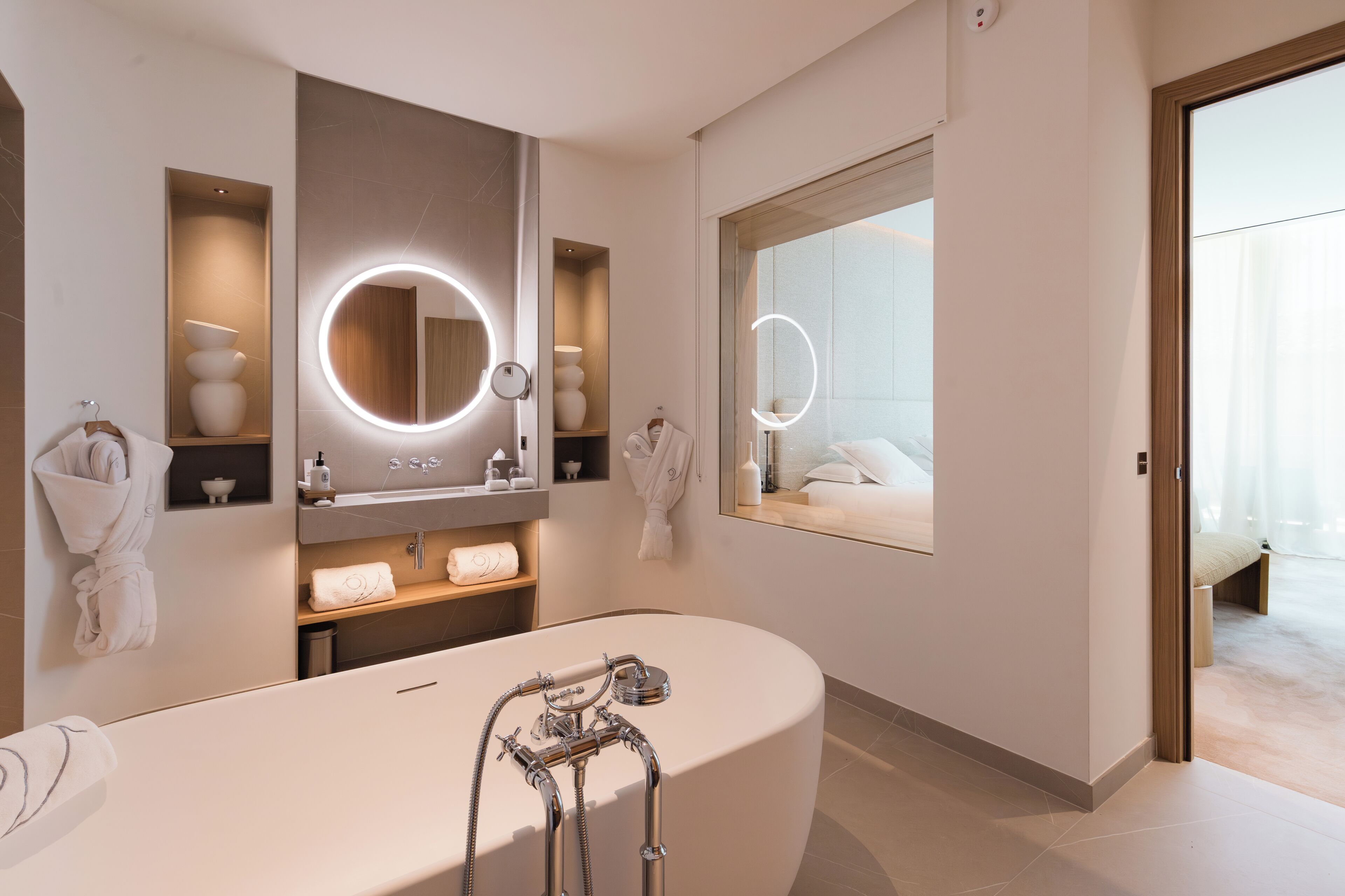 A modern bathroom with a freestanding bathtub, built-in shelves with toiletries, and a large mirror with integrated lighting.